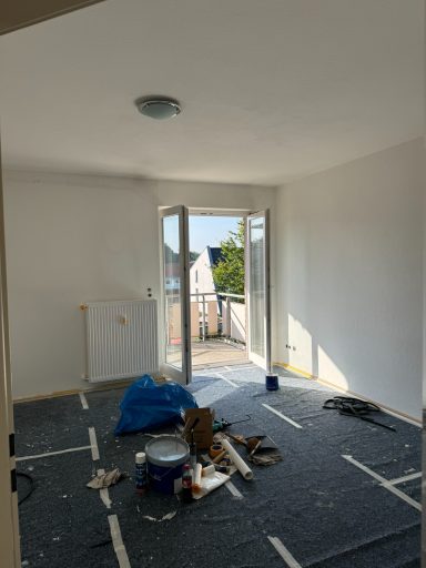 Leeres Zimmer mit offener Tür zum Balkon, Baustellenmaterial und weißer Wandfarbe.