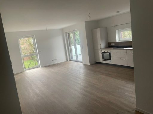 Heller, leerer Wohnraum mit Holzboden und offener Küche. Große Fenster für natürliches Licht.