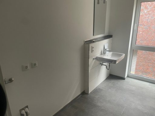 Leerer Raum mit Waschbecken, Spiegel und Fenster, grauem Bodenbelag.